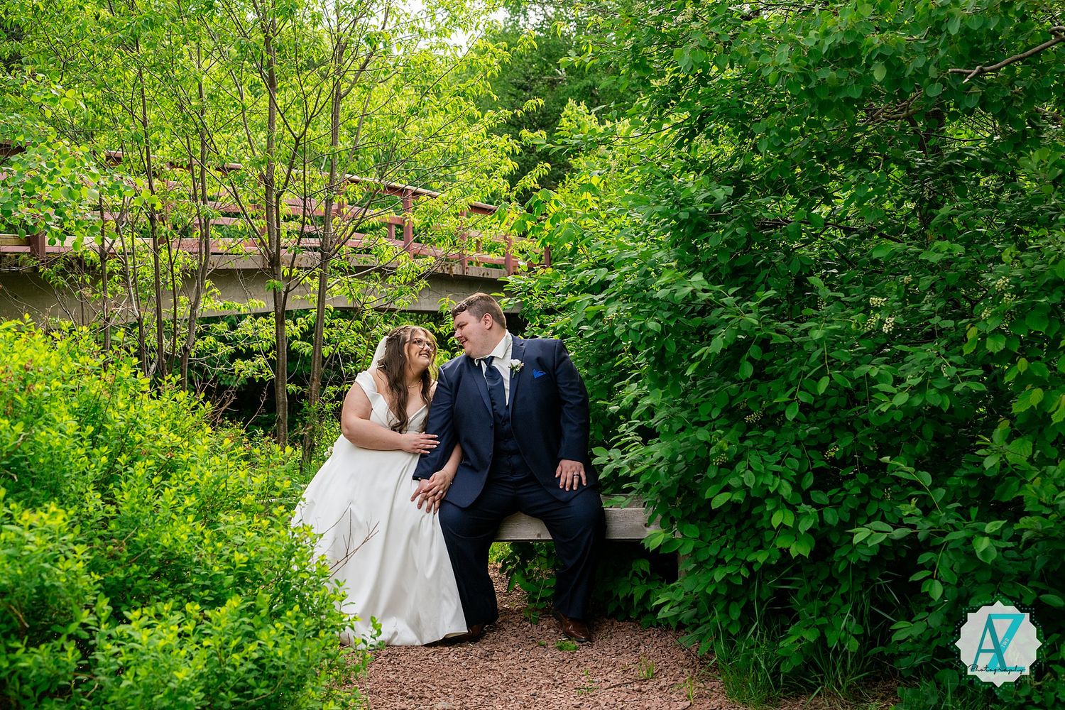 A newly married couple sits on a garden bench surrounded by lush green foliage along a winding path.