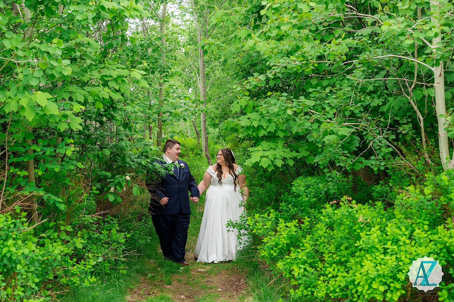 A couple walks down a lush green wooded path surrounded by vibrant summer foliage and sunlight.