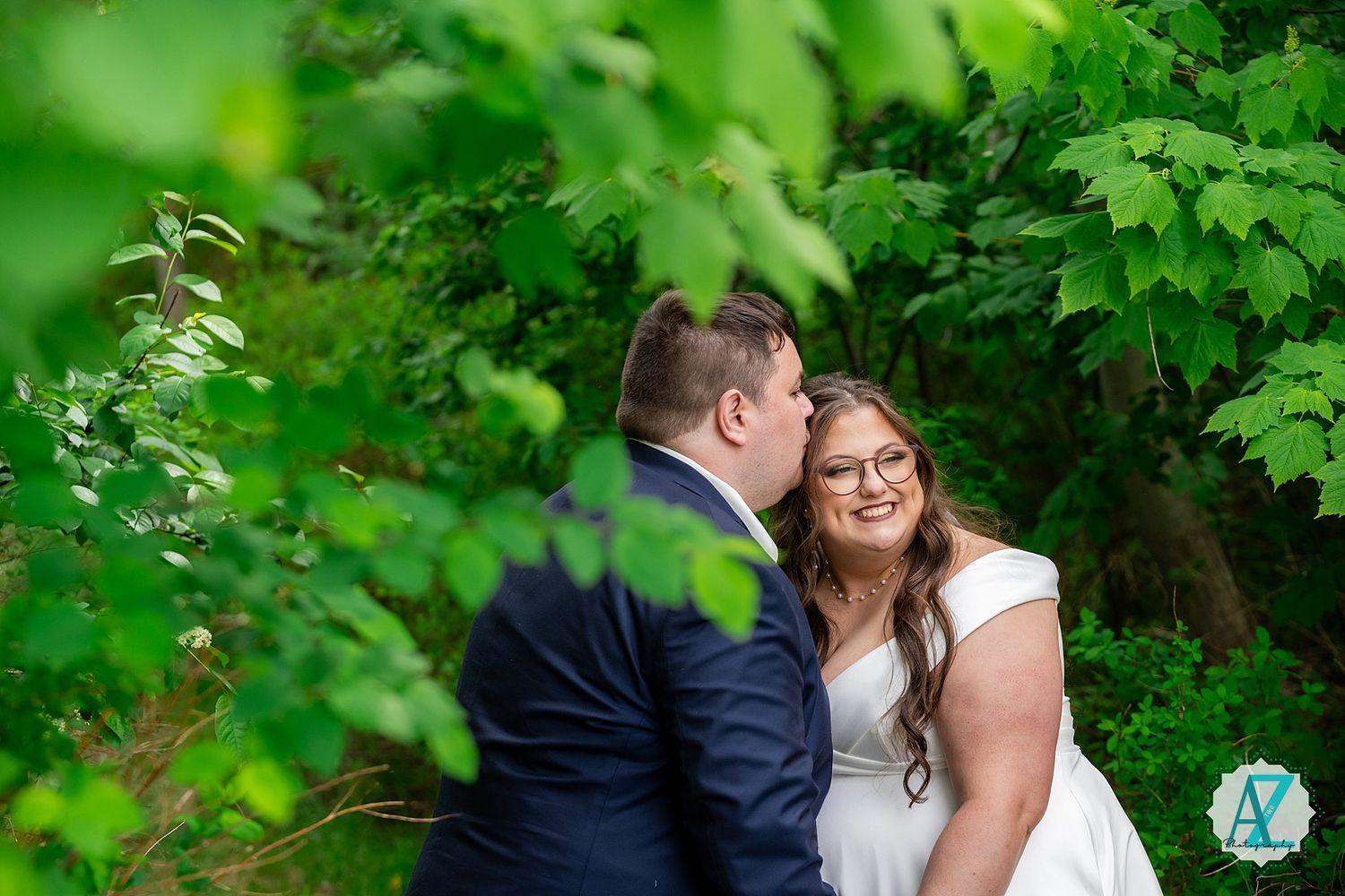 A romantic outdoor wedding photo shows a couple sharing an intimate moment among lush green foliage.
