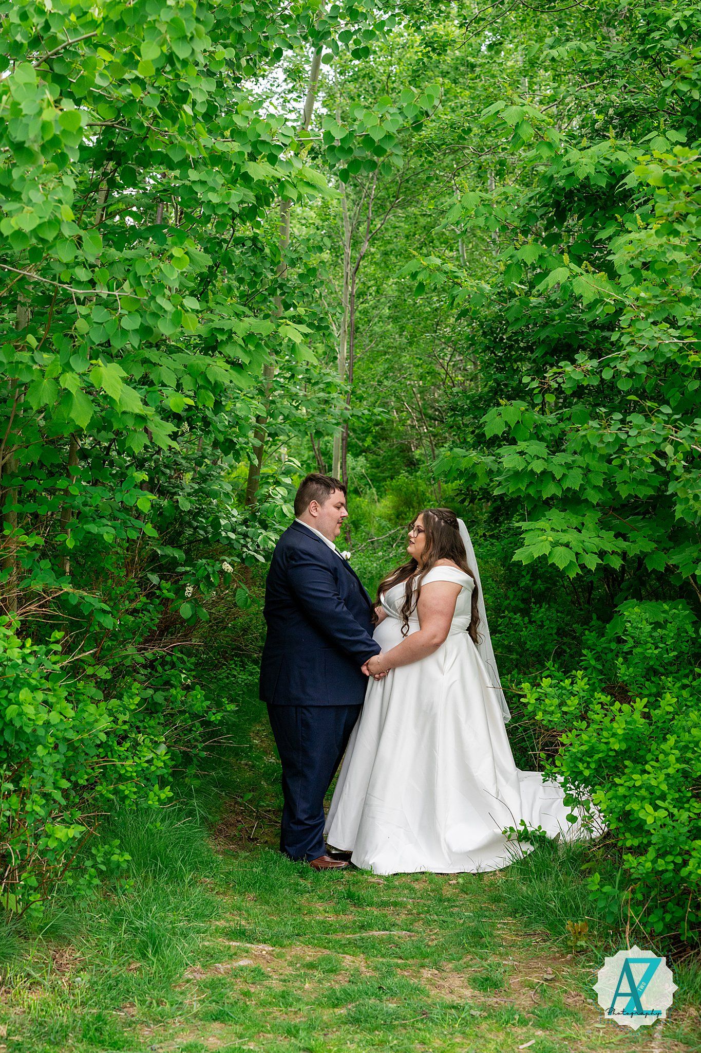 A couple embraces on a wooded path surrounded by lush green foliage on their wedding day.
