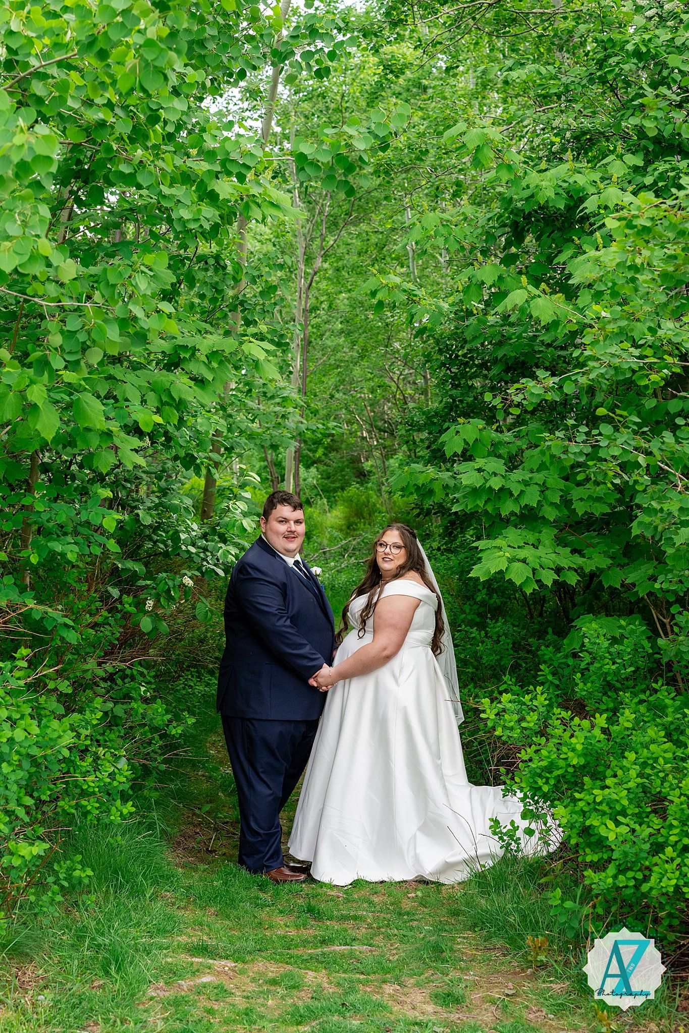 A newlywed couple poses together on a woodland path surrounded by lush green foliage on their wedding day.