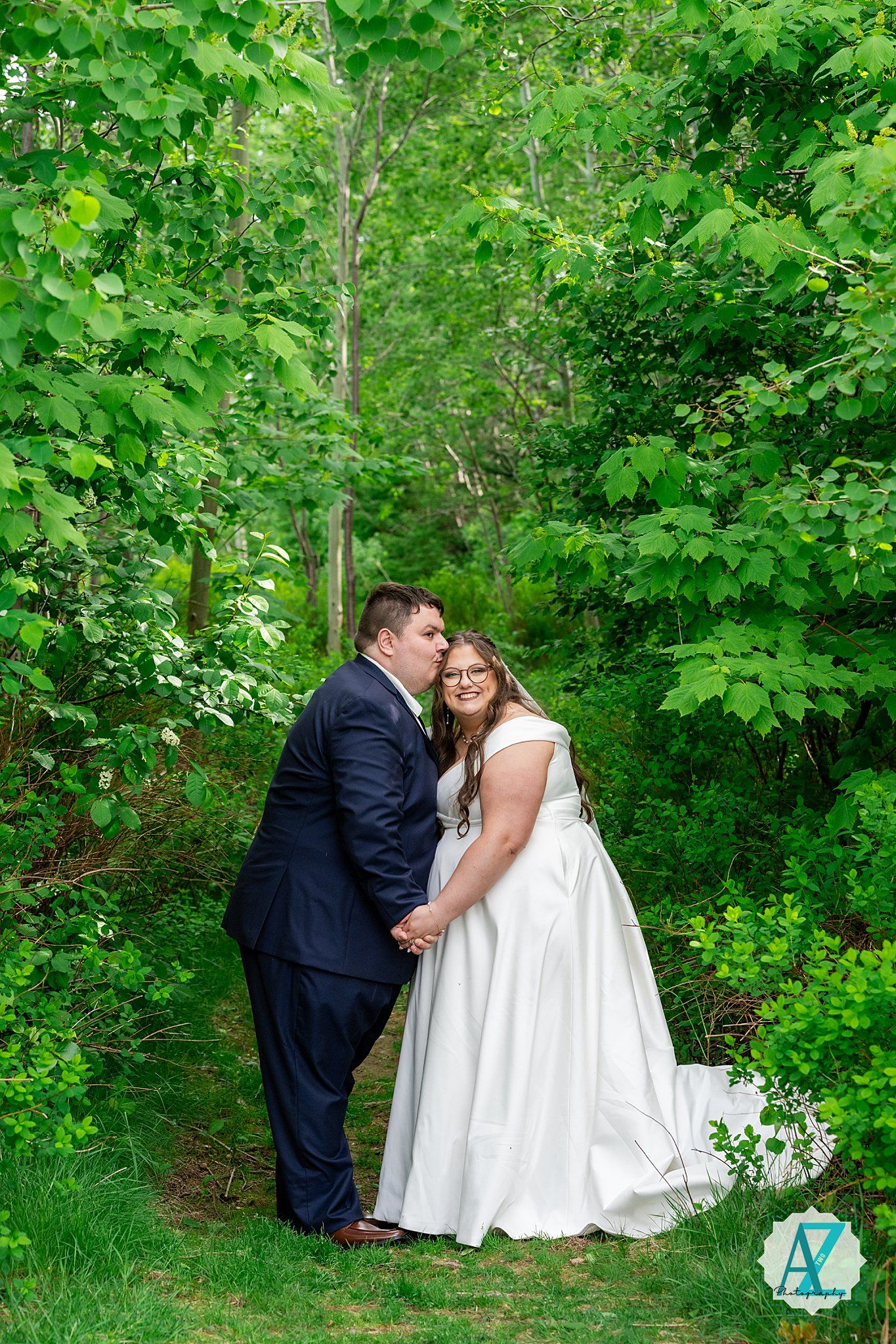 Couple in formal wedding attire share an intimate moment together on a wooded path surrounded by lush green foliage.