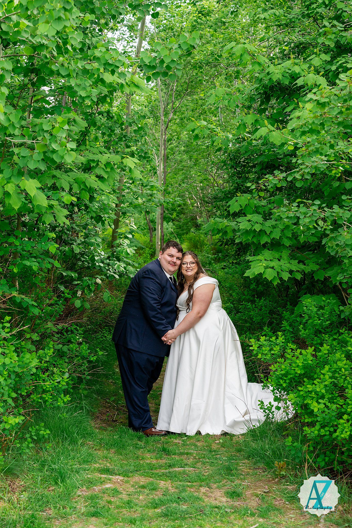 A couple in wedding attire stands on a woodland path surrounded by lush green foliage on their special day.