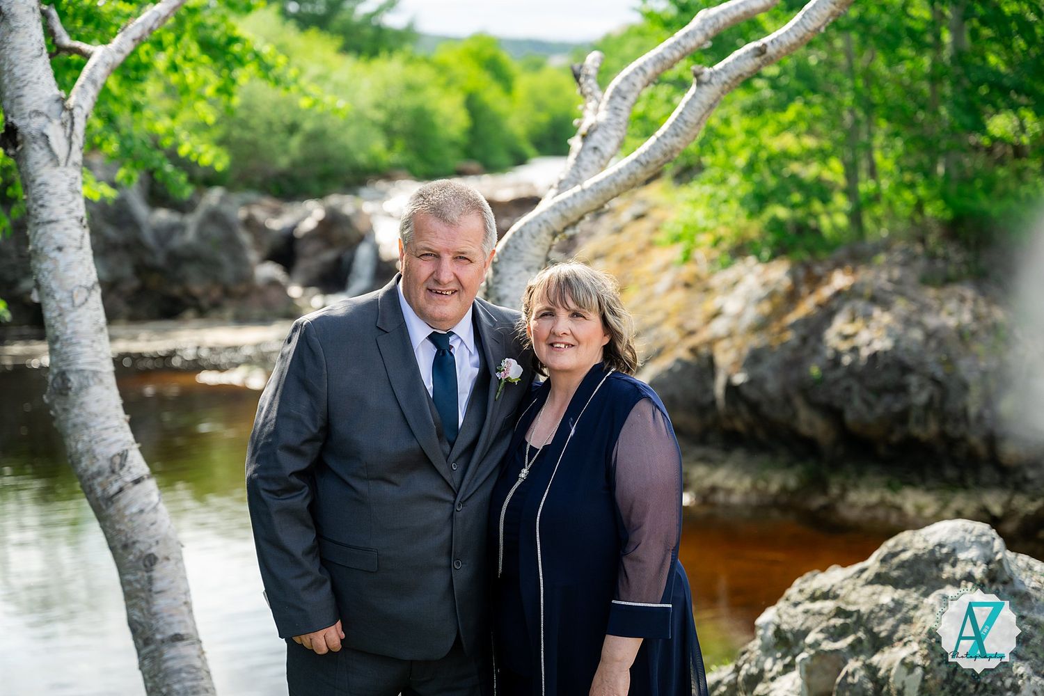 A couple in formal black attire poses for photos by a scenic waterfront with rocks and trees in the background.