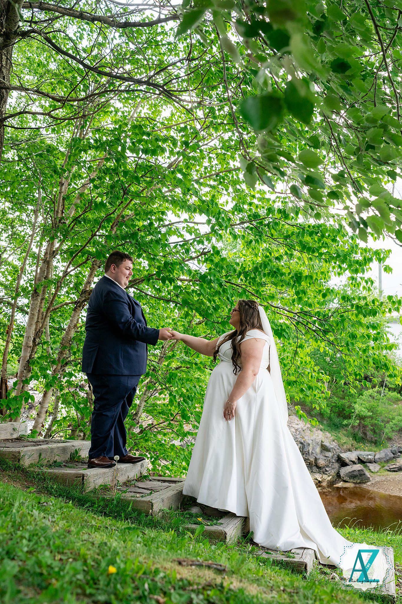 Two people hold hands while standing on stone steps surrounded by lush green foliage in an outdoor wedding setting.