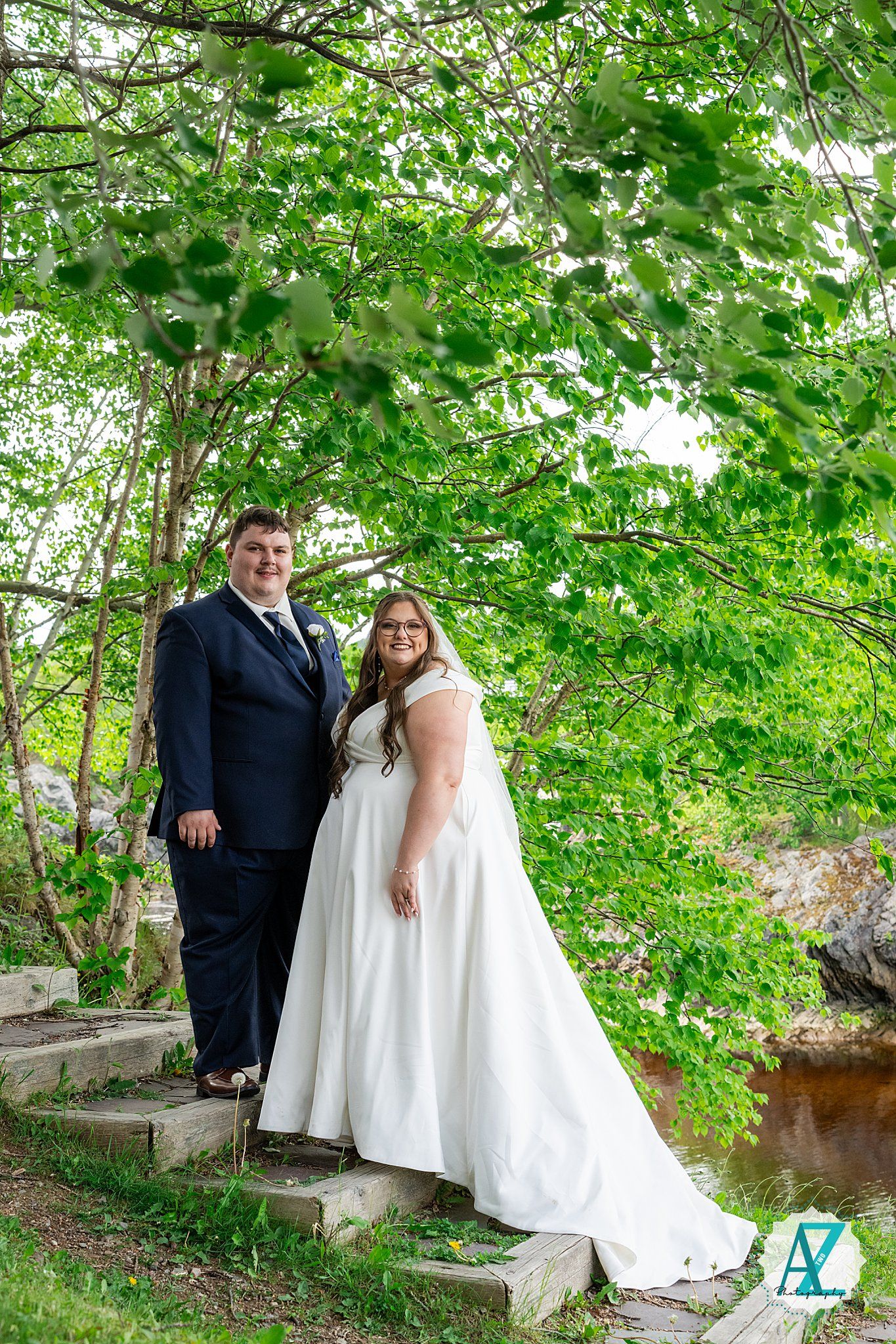 Two people share an intimate moment beneath lush green trees in a tranquil garden setting.