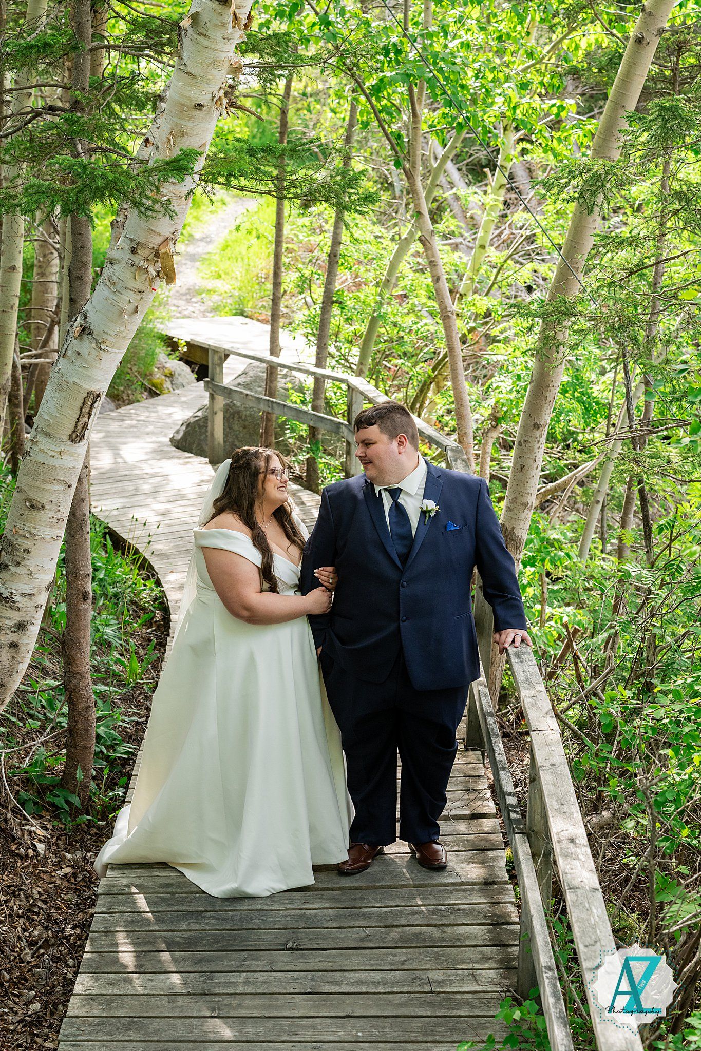 Newlywed couple walking together on a wooden boardwalk surrounded by lush birch trees and greenery.