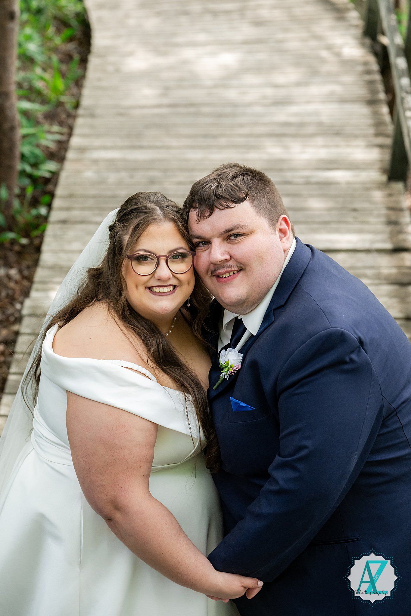 A couple in wedding attire stand close together on a wooden walkway surrounded by greenery.