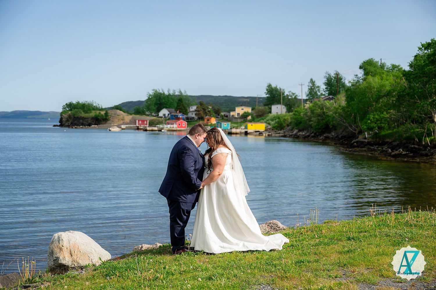 A couple shares a romantic moment on grassy shoreline overlooking scenic lake waters with boats and trees in background.