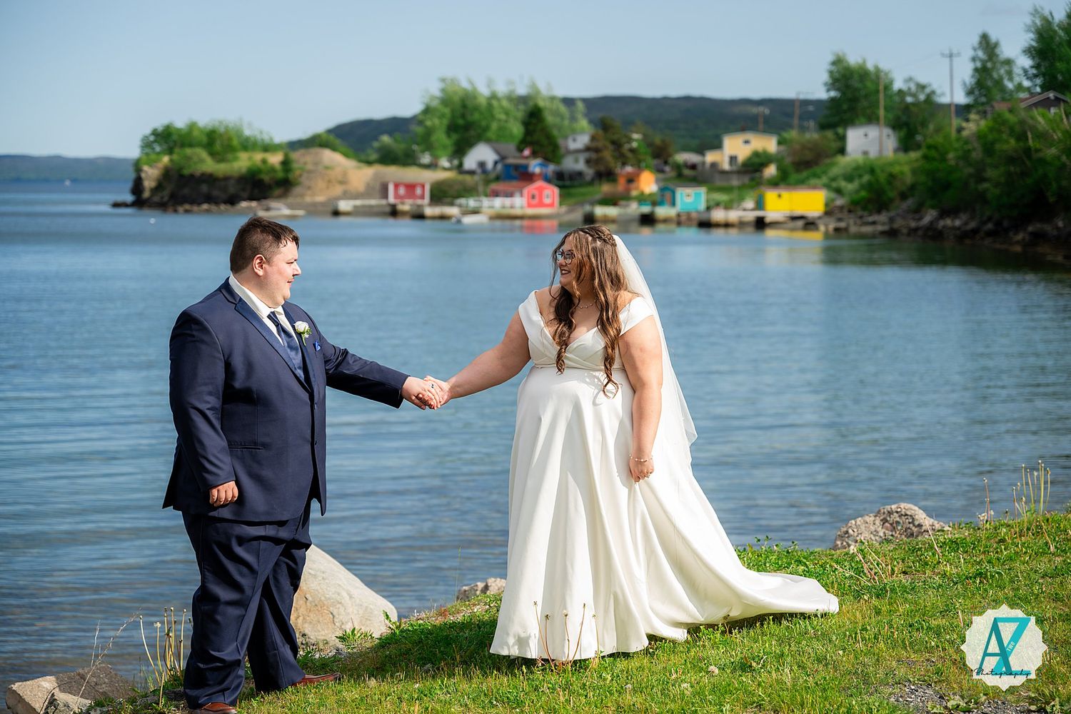 Wedding couple sits by scenic waterfront with colorful buildings and boats in background.
