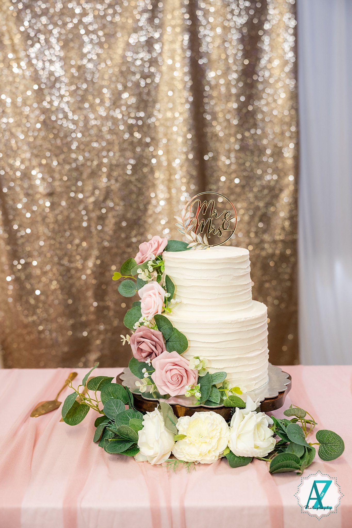 Three white wedding cakes decorated with pink roses and eucalyptus greenery sit on a table with gold sparkly backdrop.