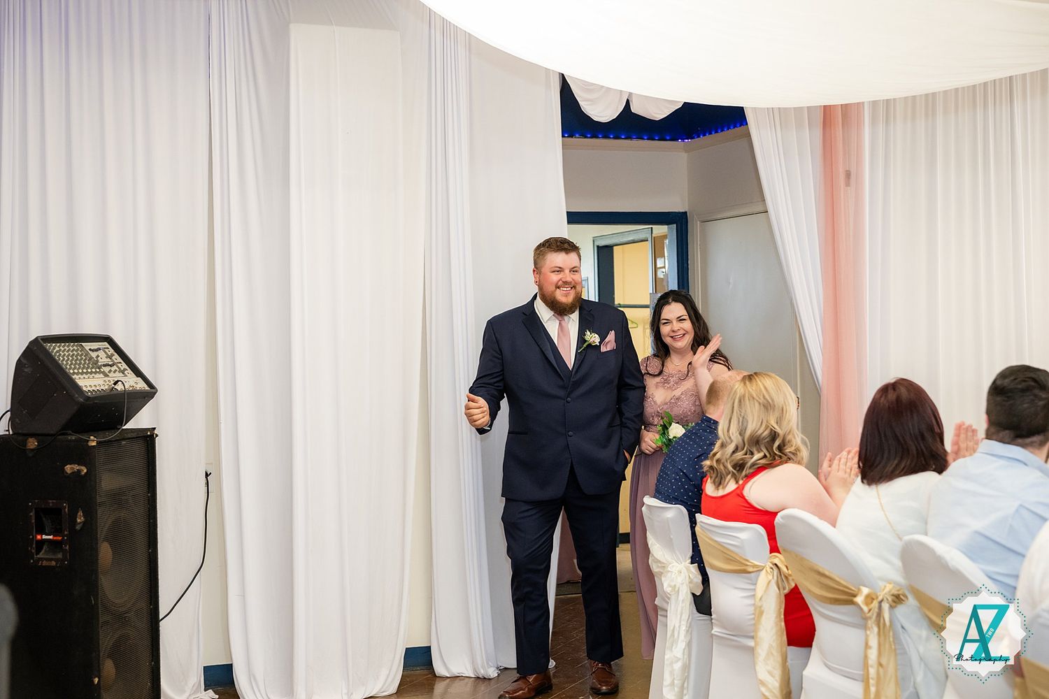 A wedding reception venue decorated with white curtains shows guests mingling and dancing at the celebration.