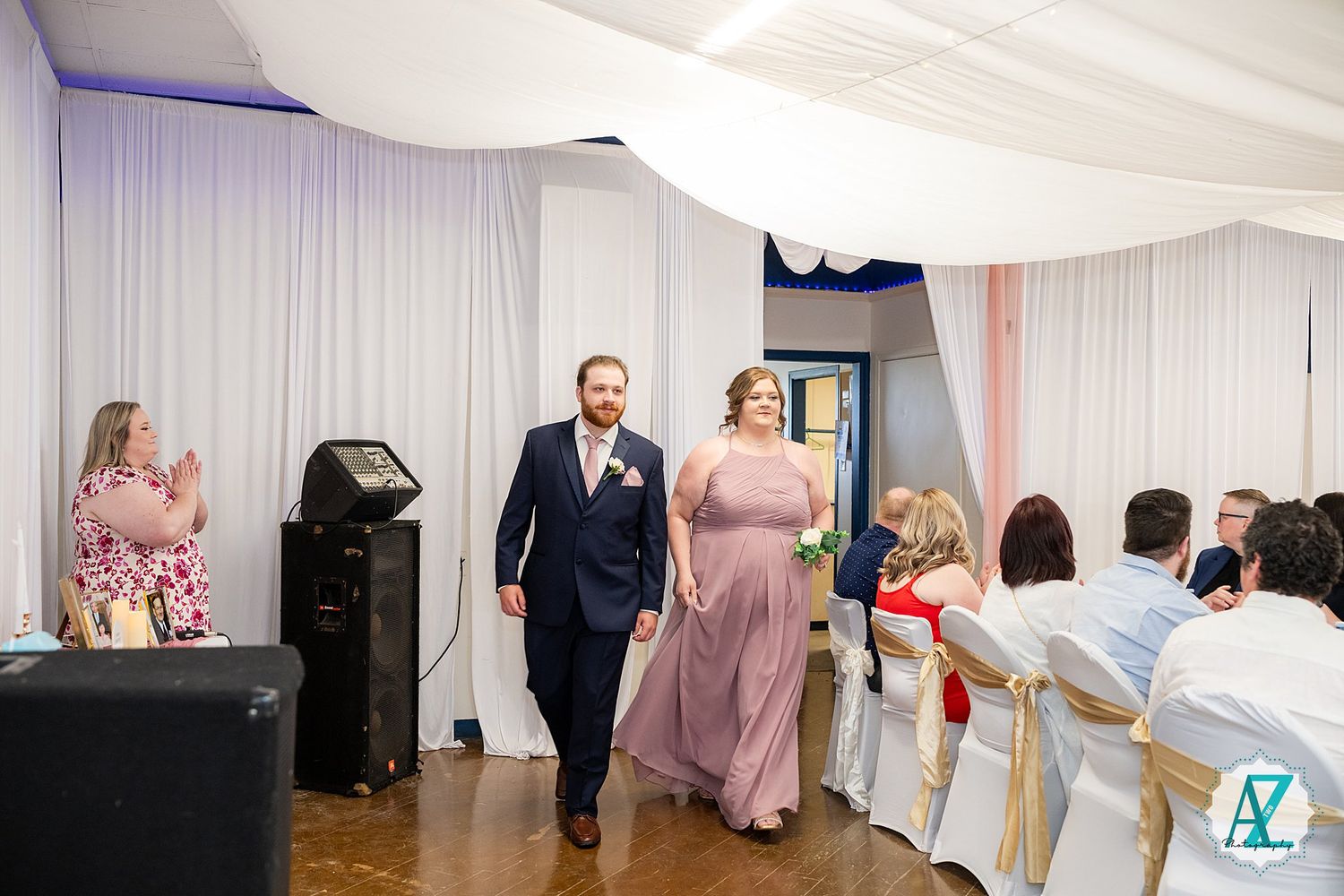 Members of a wedding party enter a reception venue decorated with white drapes and chiavari chairs at a formal event.