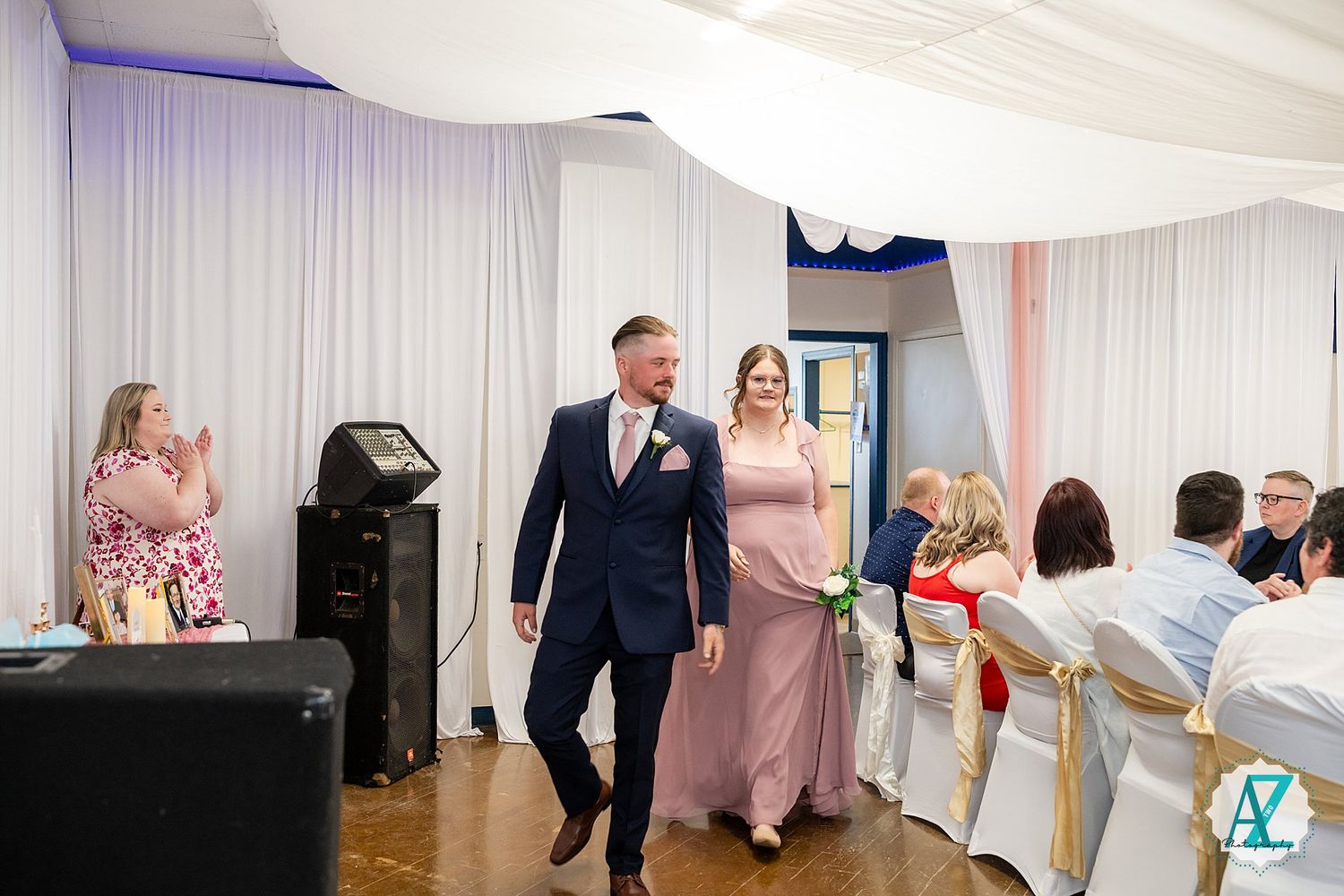 Wedding guests applaud as a couple in navy and pink formal attire make their entrance at a reception with white draping.
