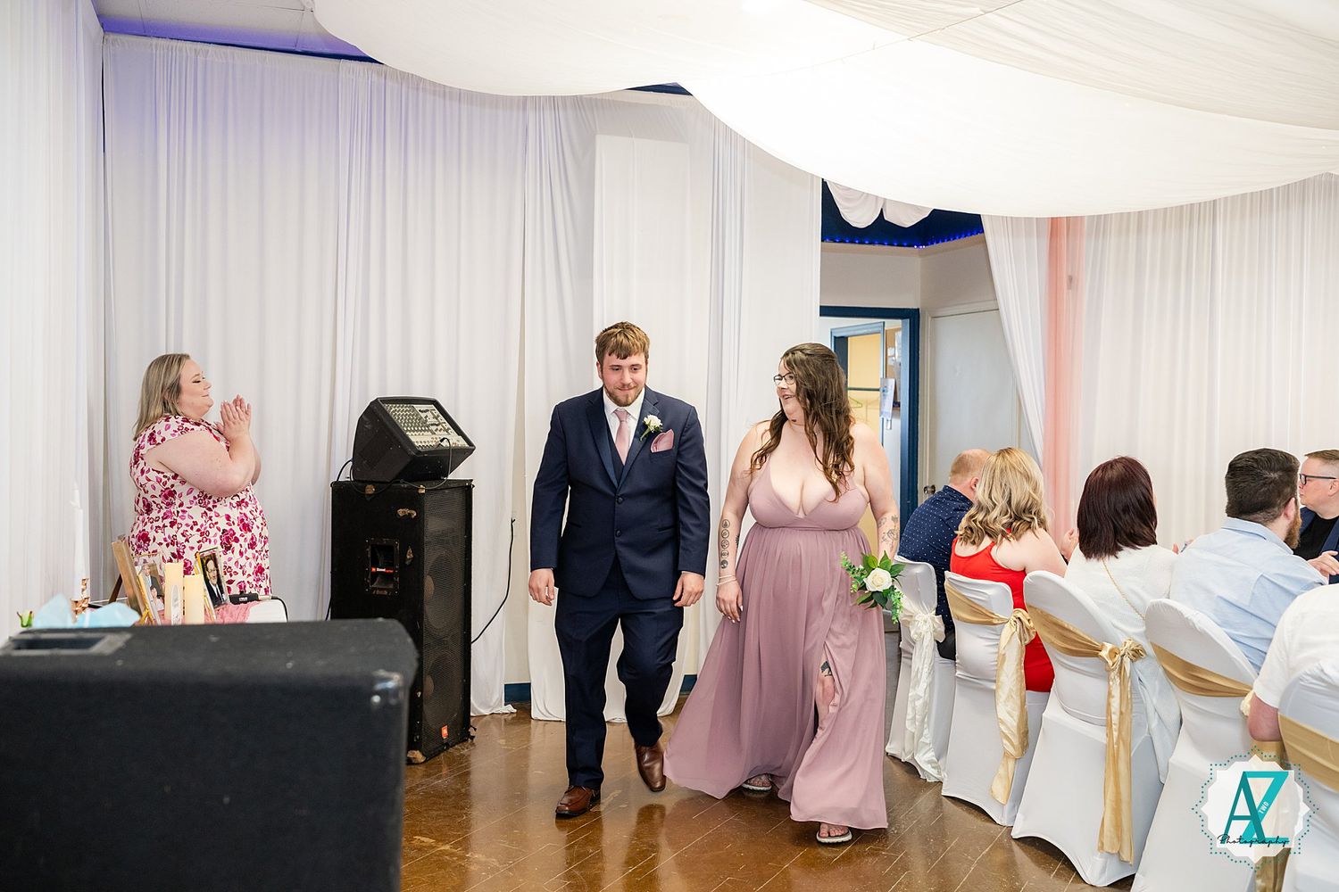 Wedding party members in formal attire make their entrance at an indoor reception venue with white draped walls.