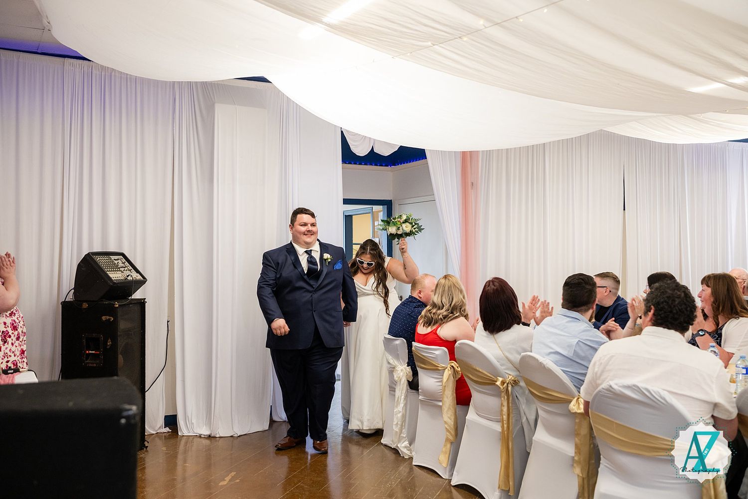 Couple makes a grand entrance at wedding reception as guests sit at decorated white tables with curtained backdrop.