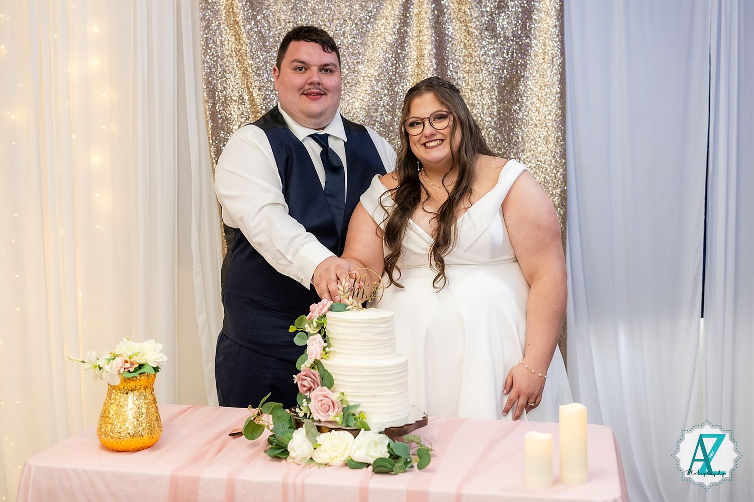 A couple stands behind their elegant white wedding cake decorated with fresh flowers at their reception.
