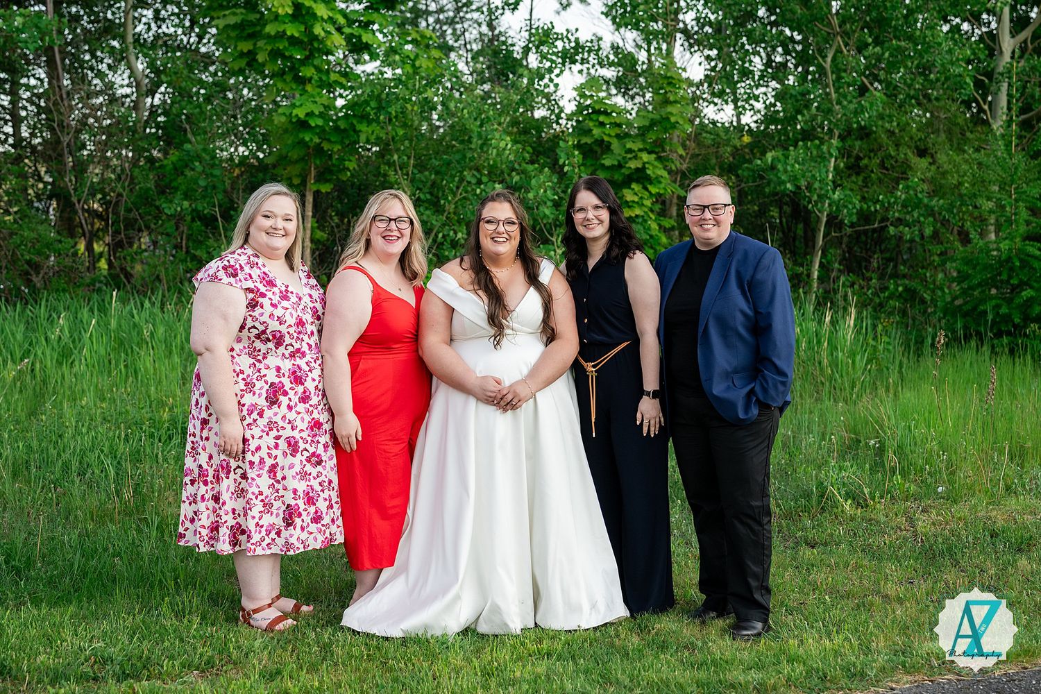 A group of people wearing formal attire pose together in an outdoor setting with lush green trees in the background.