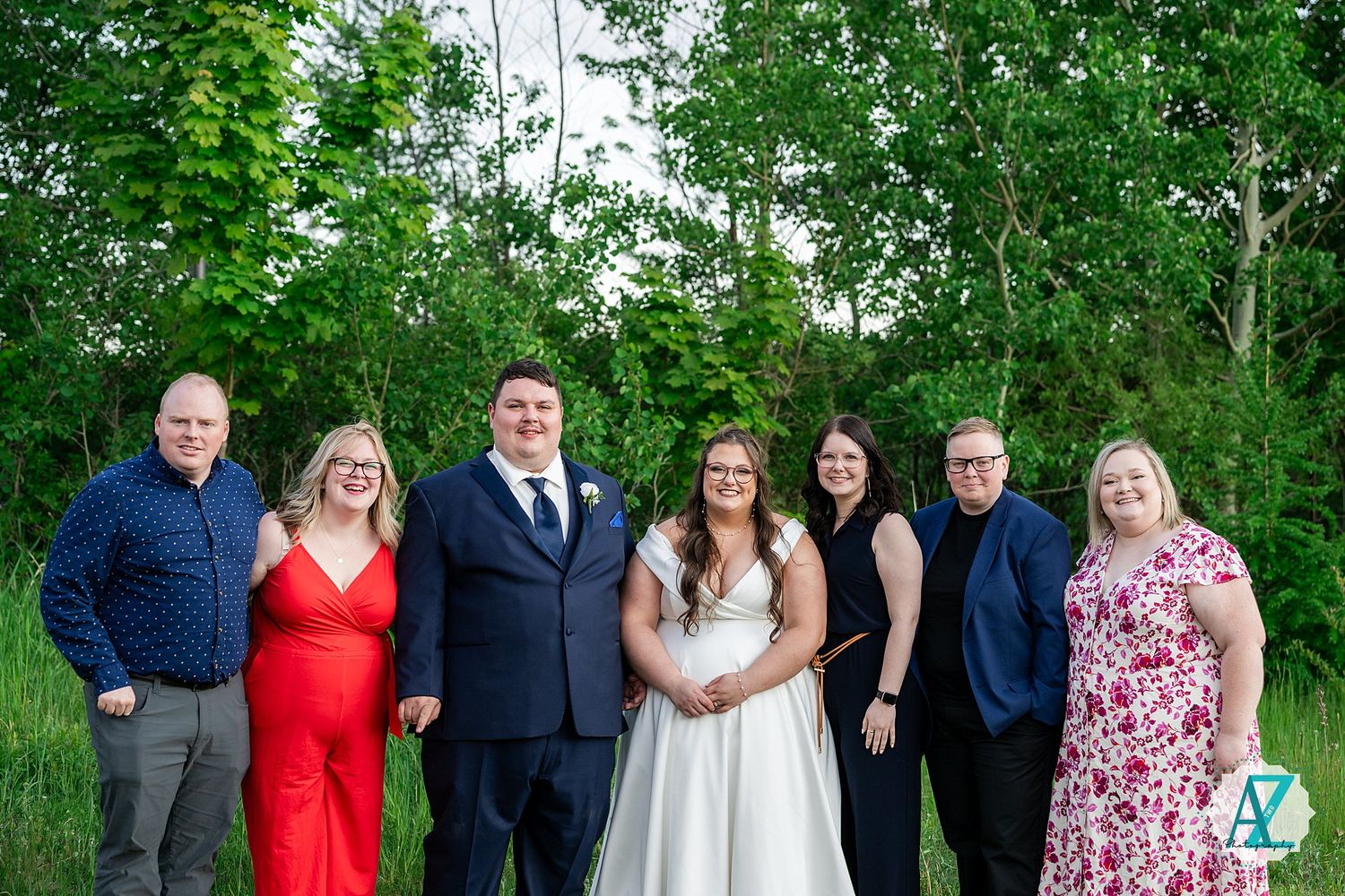 A wedding party stands in line against a backdrop of lush green trees wearing formal attire in blue and red.