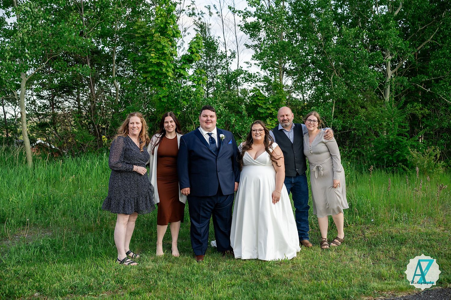 A small wedding party poses together on lush green grass with trees in the background during a summer celebration.