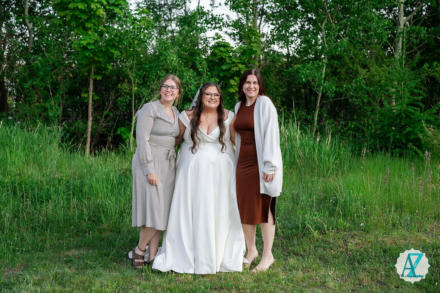 Three people in light colored dresses stand together on lush green grass with trees in the background.