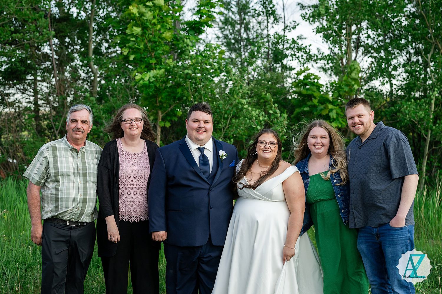 Group gathered outdoors for a wedding celebration standing together against lush green trees and foliage in summer.