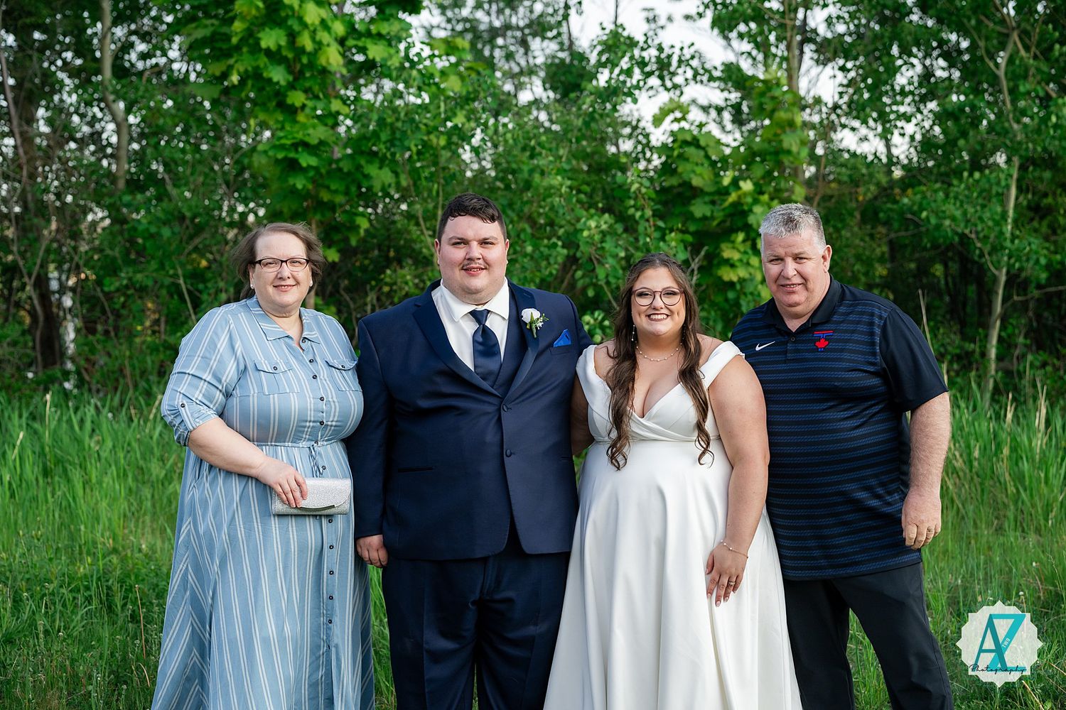A group of people in formal attire pose together outdoors against a backdrop of lush green trees and foliage.