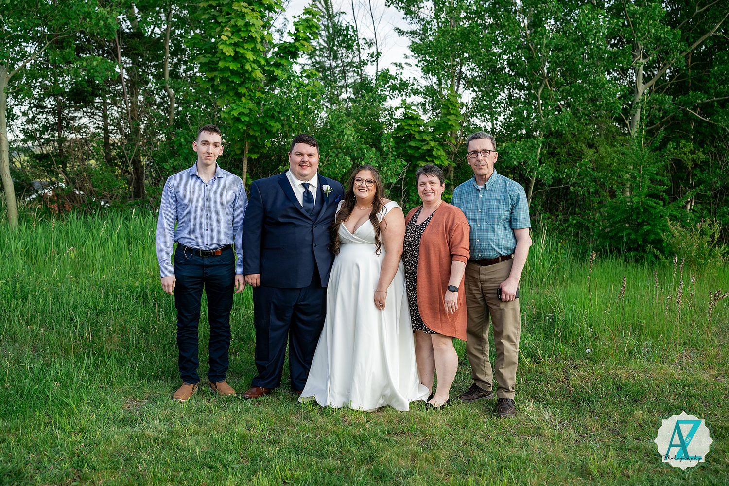 A group stands together on green grass with trees in the background at an outdoor wedding celebration.