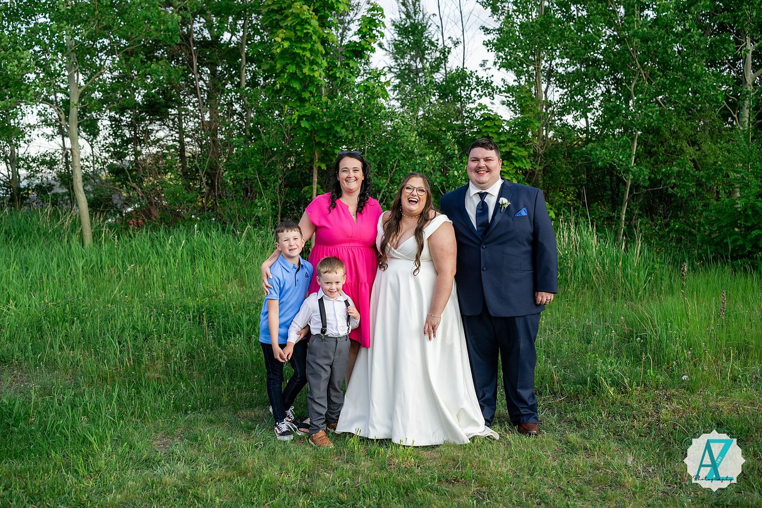 Group of people in formal attire pose together for outdoor portrait in front of green wooded backdrop.
