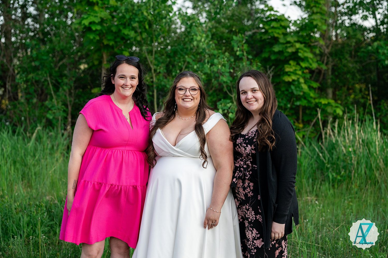 Three friends pose together outdoors against a backdrop of tall green grass and trees on a summer day.