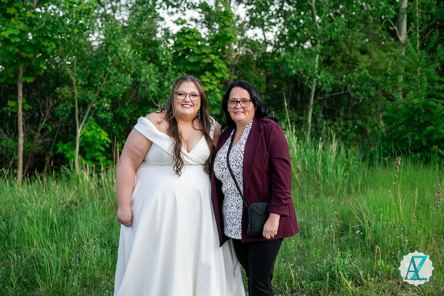 Two people pose together in a grassy meadow with trees in the background during an outdoor photo session.