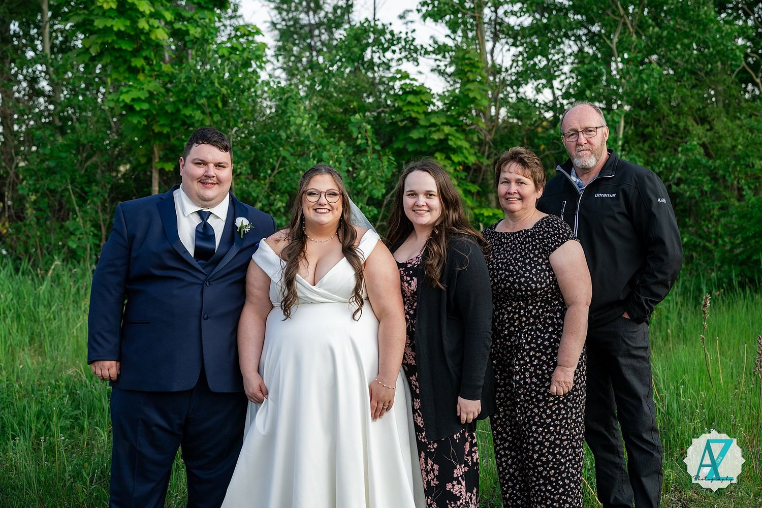 Wedding party poses outdoors in front of lush green foliage on a sunny summer day.