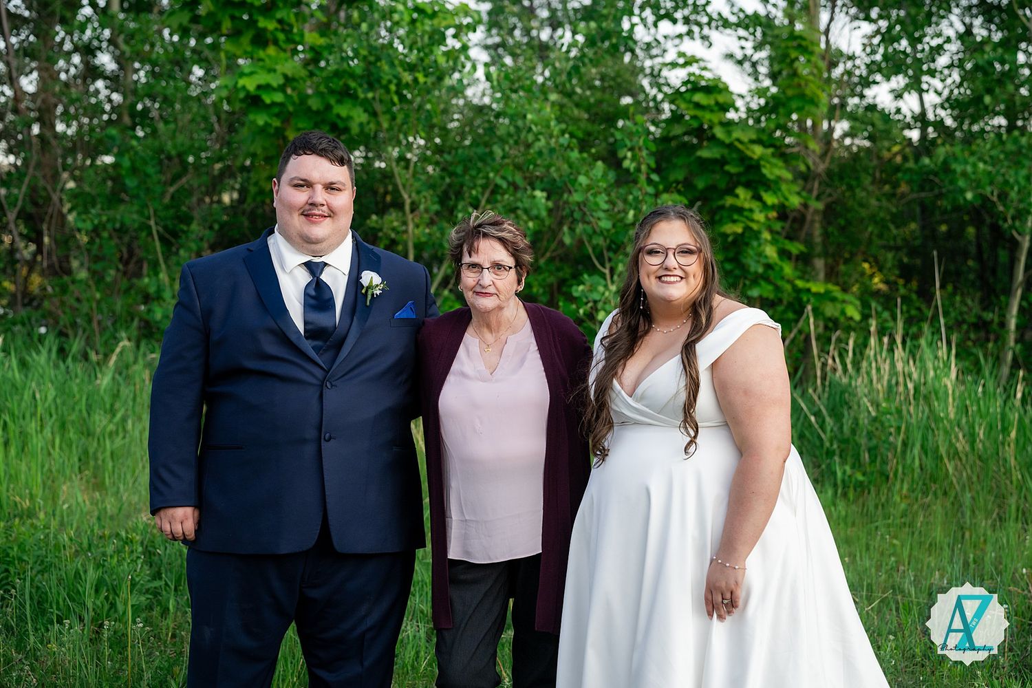 Three people stand together outdoors in formal attire with green foliage in the background.
