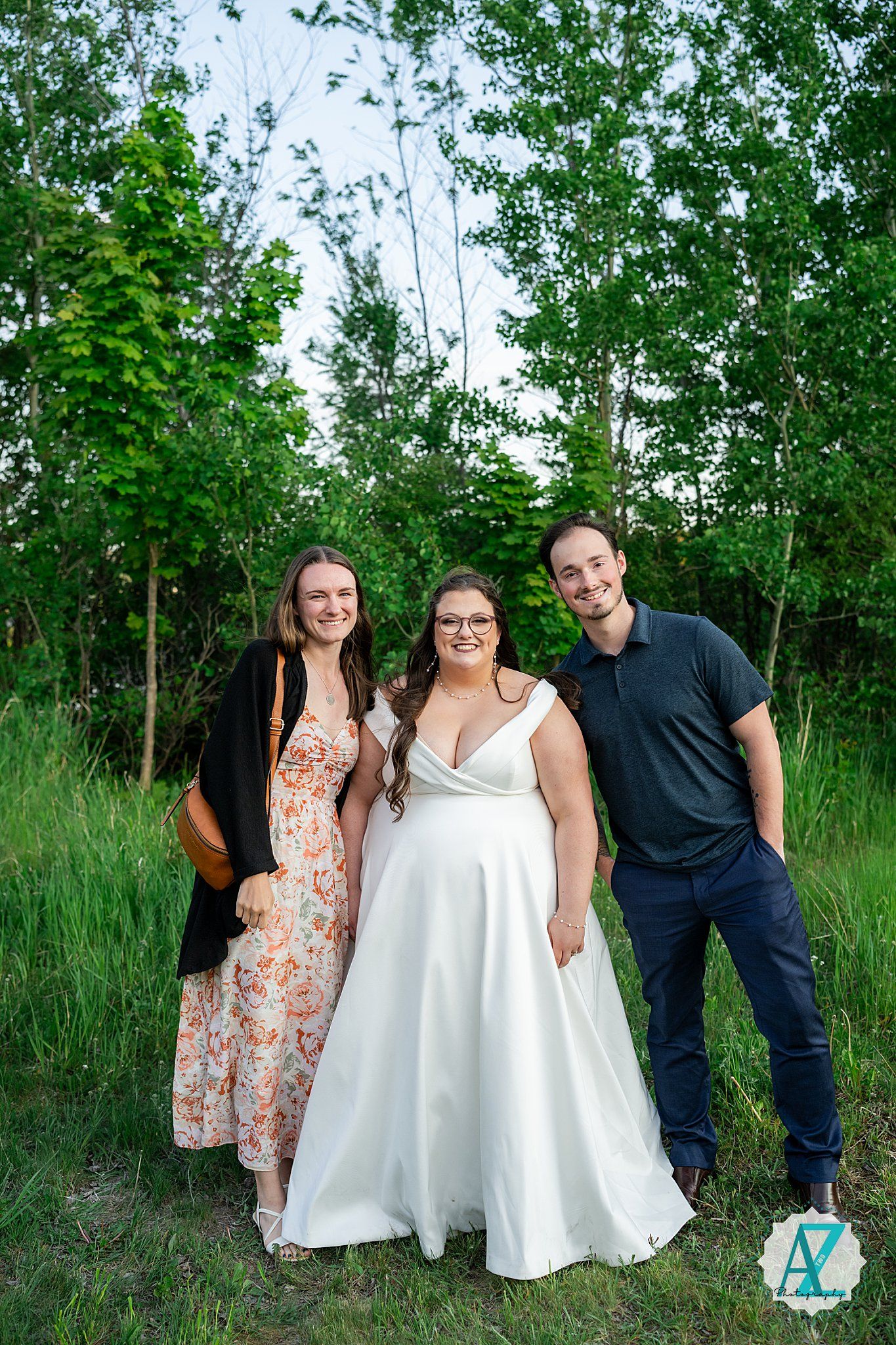 A group poses playfully together outdoors in formal attire against a backdrop of green summer foliage.