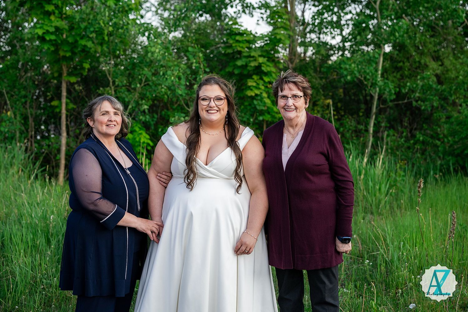 Three figures in formal attire stand together in a natural outdoor setting with green trees and tall grass.