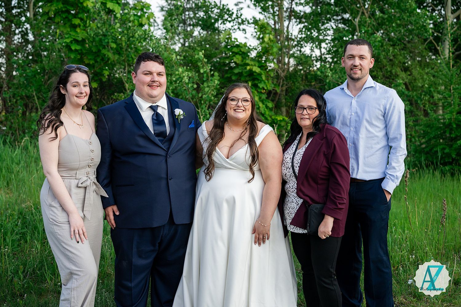 A group of wedding guests stands together in a green field with trees in the background for an outdoor photo.