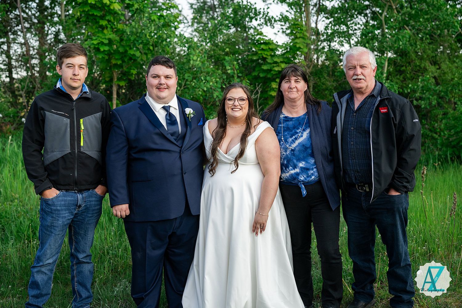 A group stands together in front of lush green foliage wearing formal attire for an outdoor photo.