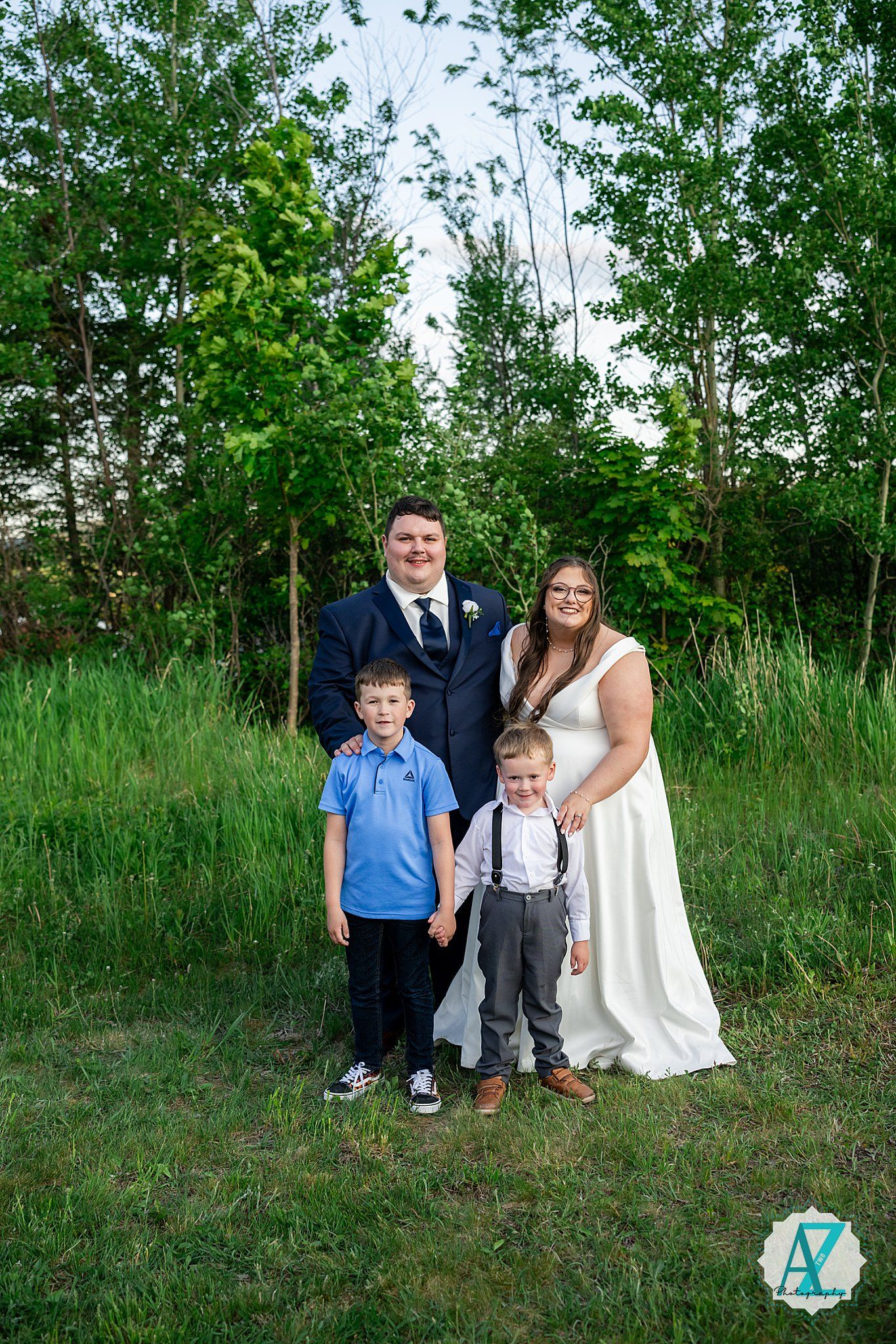 Wedding couple stands with children in a lush green meadow surrounded by tall grasses and trees on a sunny day.