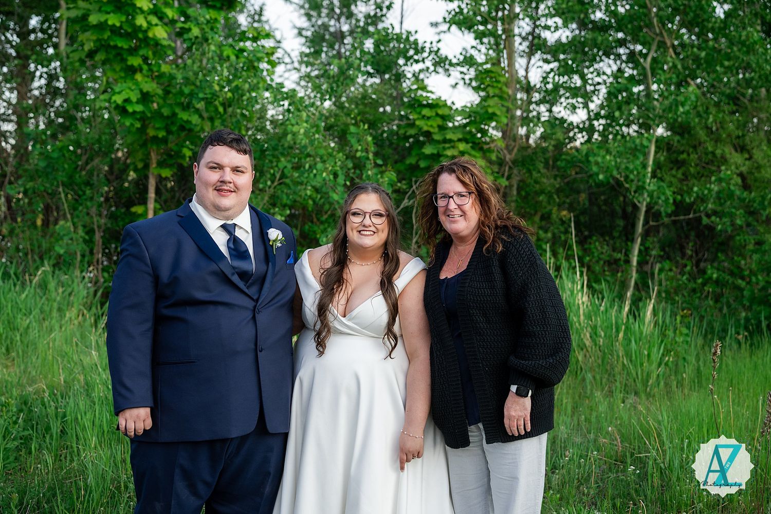 Wedding party stands together on a lush green lawn surrounded by tall trees in the background.