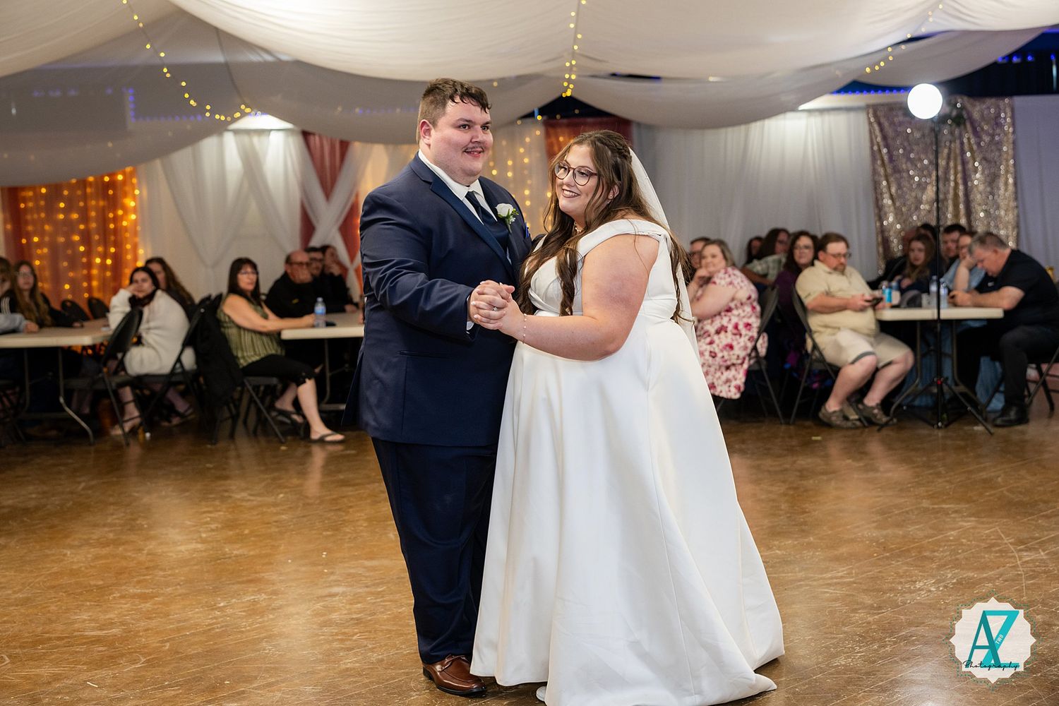 A couple dances in a slow embrace on a large reception hall floor during an elegant indoor wedding celebration.