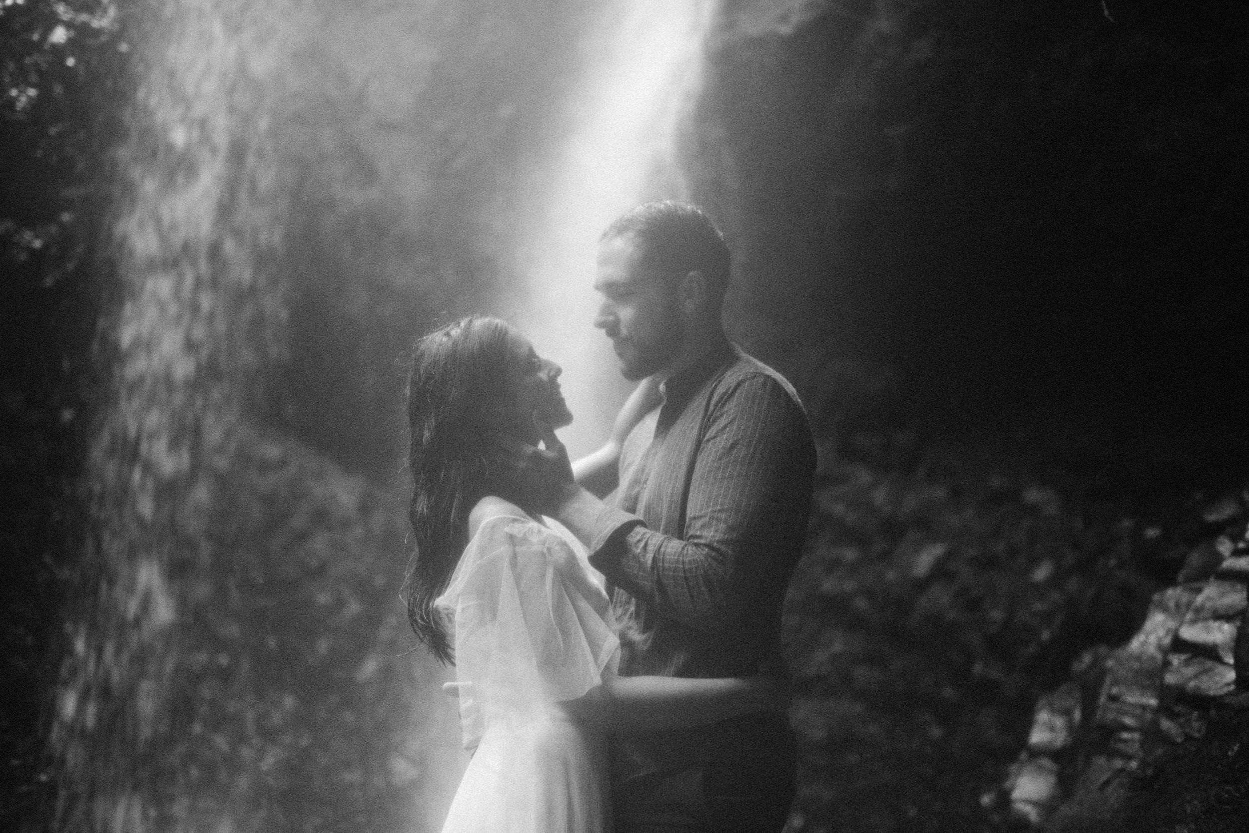 A romantic black and white moment captured under a misty waterfall showing a tender embrace.