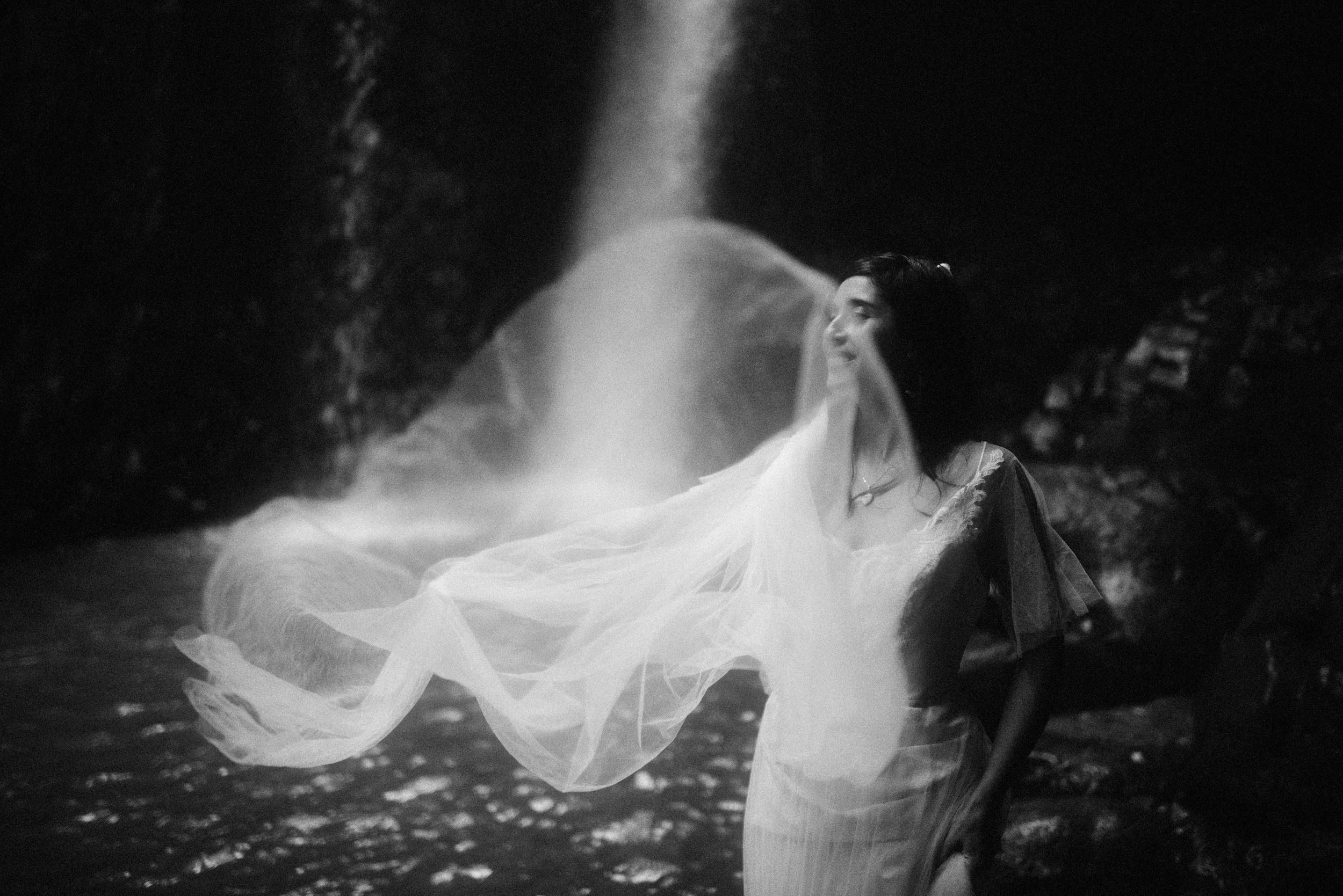 A ghostly figure in flowing white stands in front of a cascading waterfall creating an ethereal black and white scene.