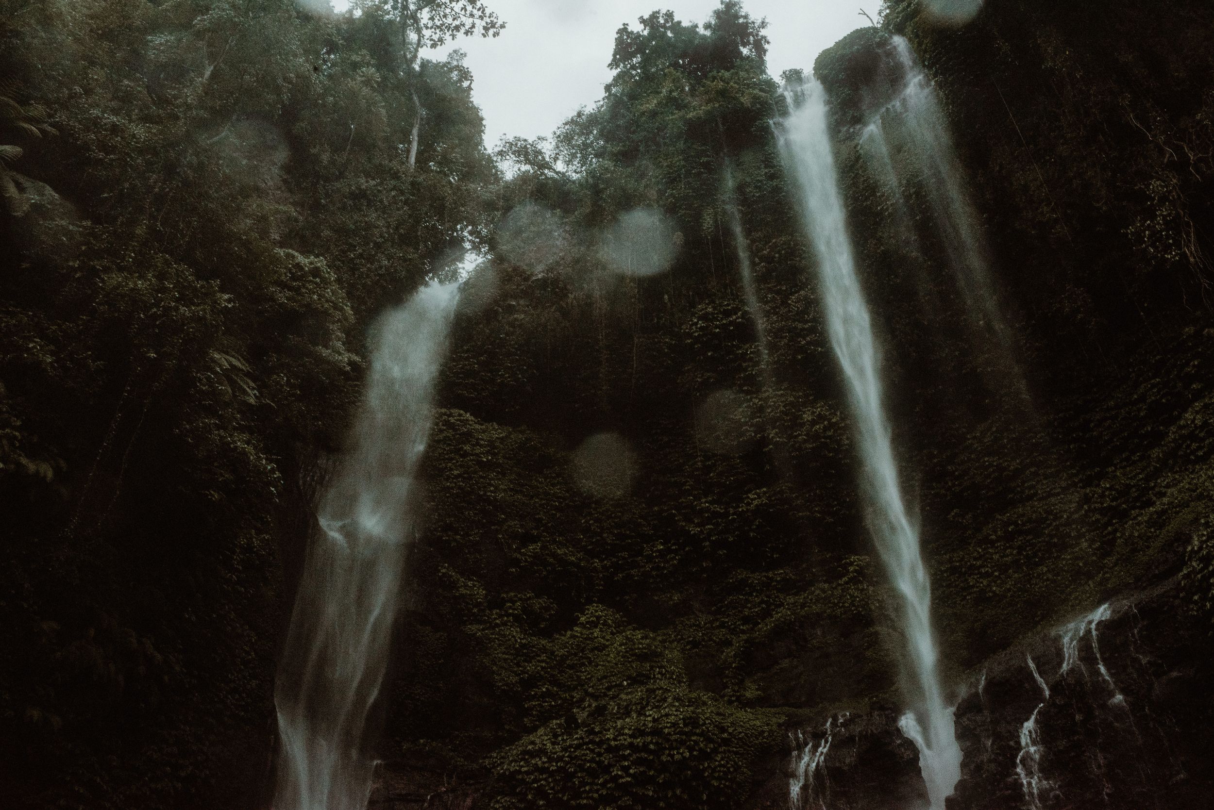 Misty twin waterfalls cascade down a dark tropical forest cliff with sunbeams streaming through the canopy.