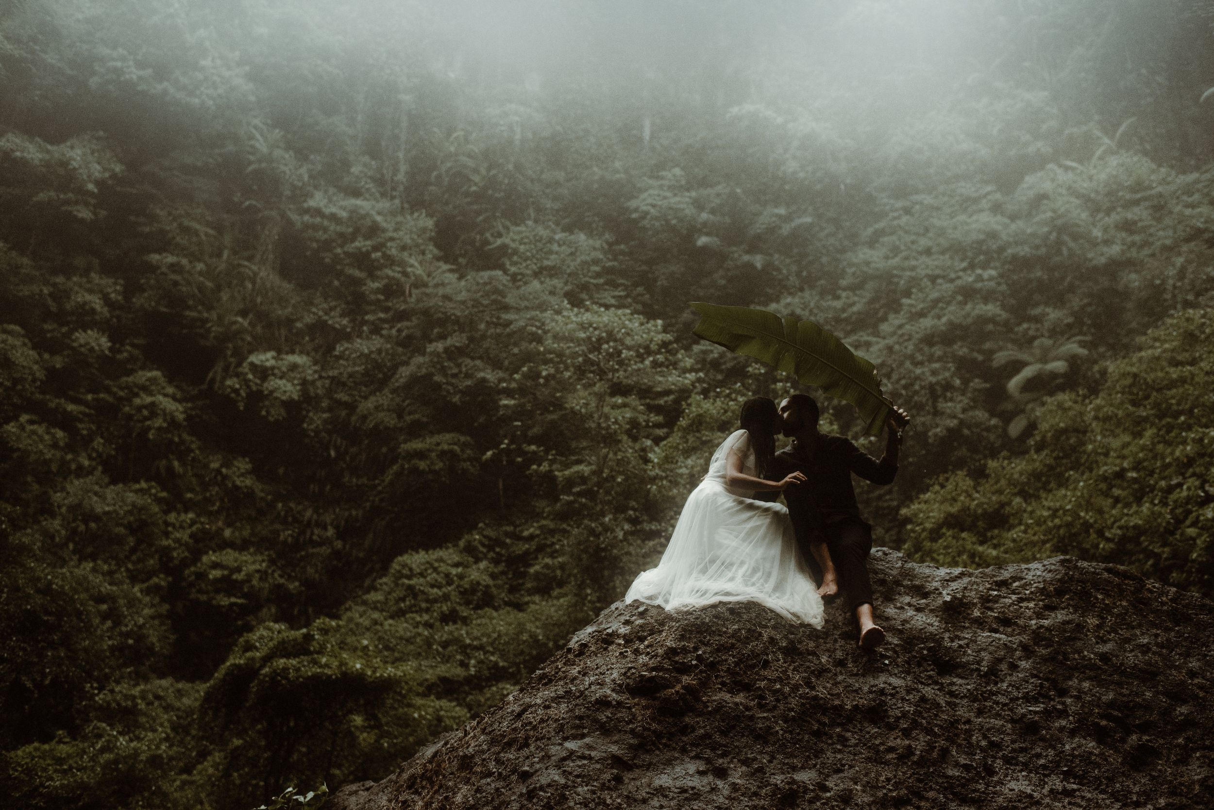 Two people in wedding attire embrace on a misty mountain ledge surrounded by lush green forest landscape.