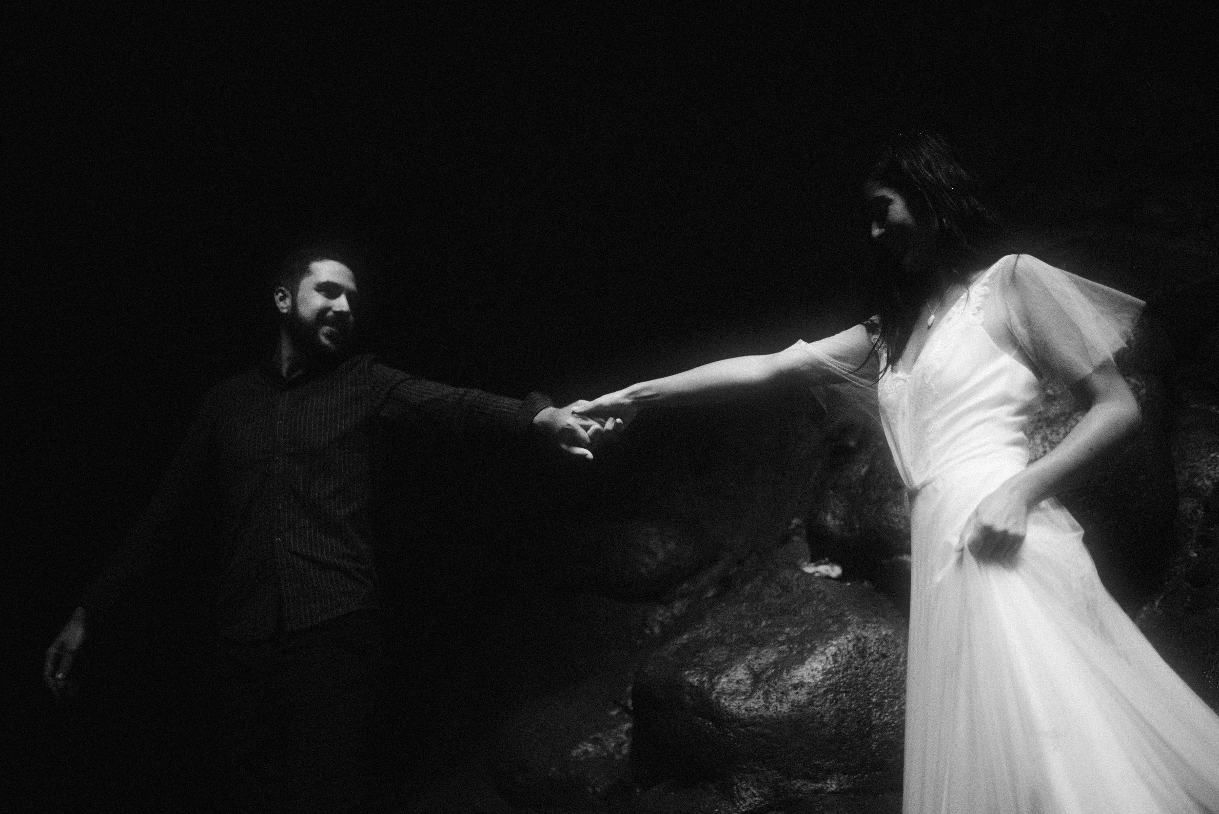 Black and white artistic photograph of two people holding hands in a dreamy outdoor setting at night with a dramatic atmosphere.