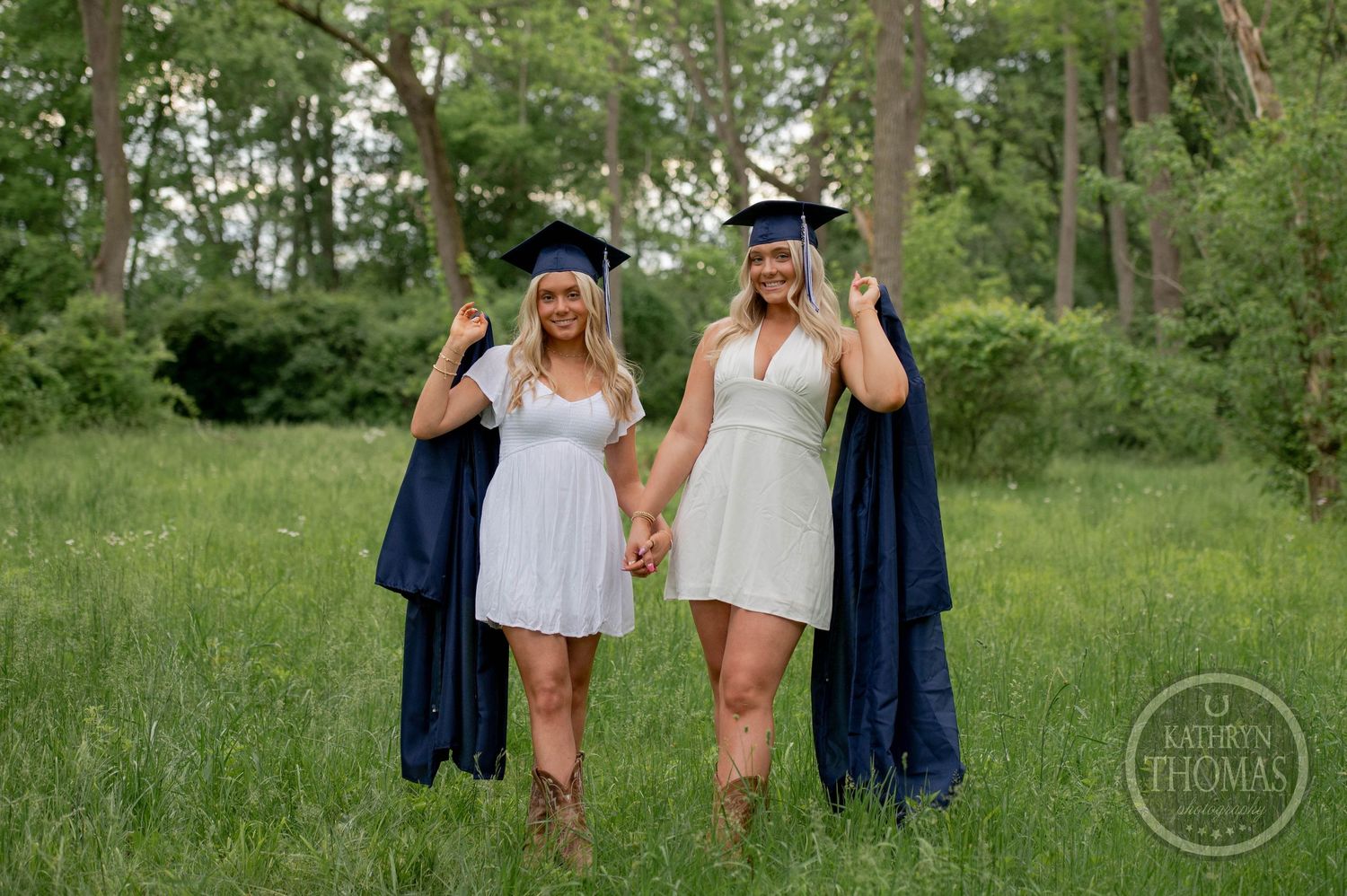 Two students in graduation attire toss their caps in celebration while standing in a lush green meadow surrounded by trees.