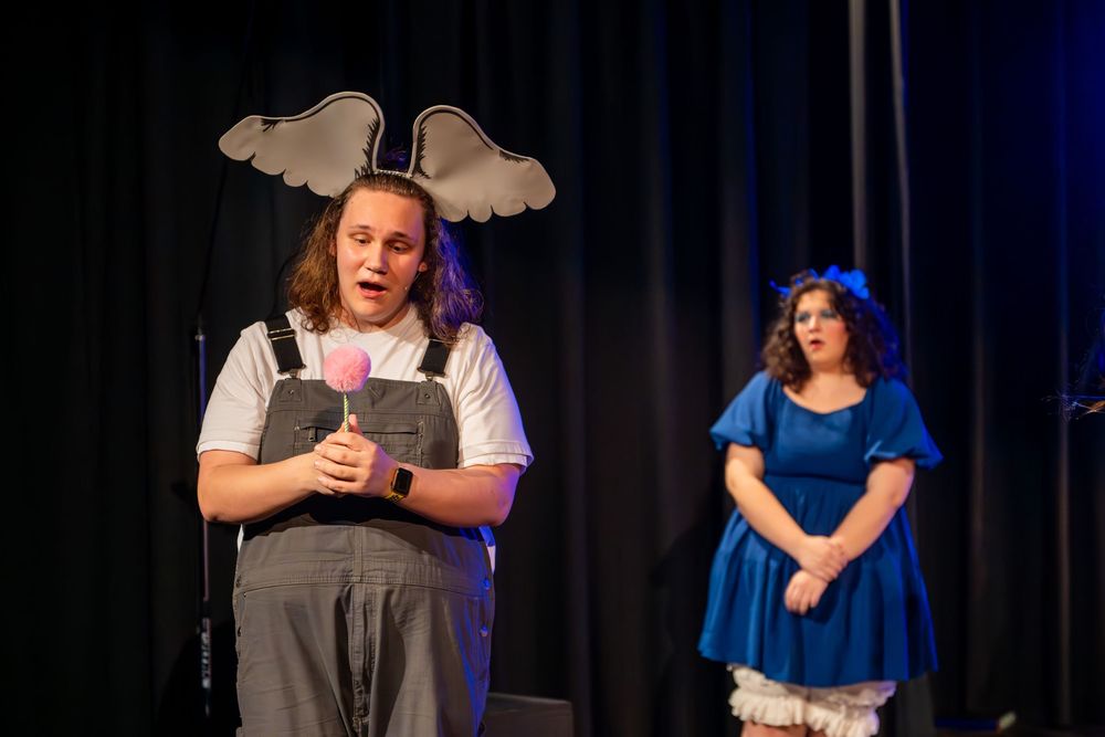 Stage performers in grey and blue outfits act in a theatrical production with a dark curtain backdrop.