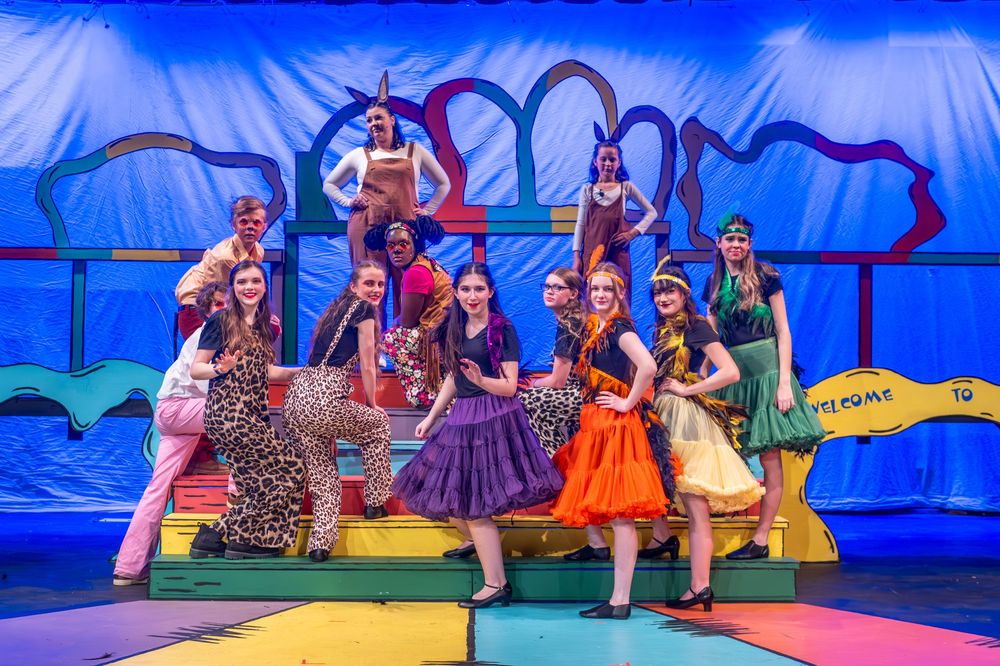 Musical theater performers in colorful 1950s-style costumes pose energetically on a vibrant stage set with heart decorations.