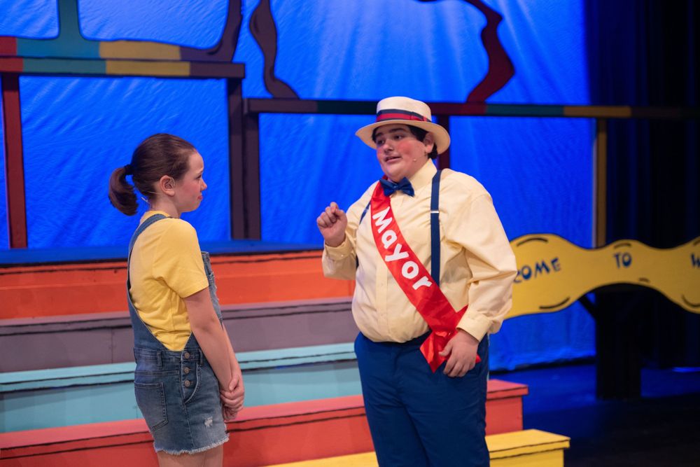 Two performers interact on a colorful stage set with one wearing a yellow shirt and the other in white with a sash.