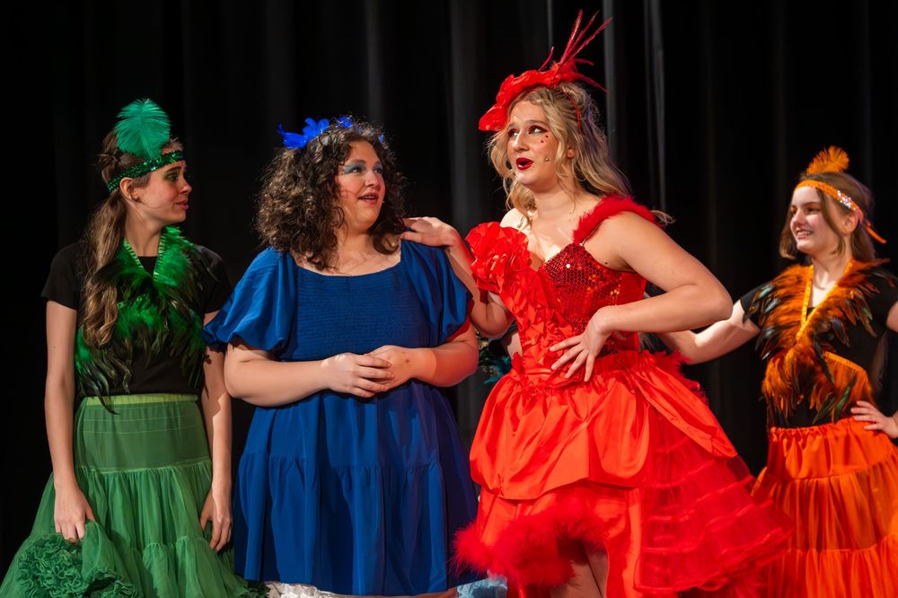 Group of performers on stage wearing colorful dresses in green, blue, red and orange during a theatrical production.